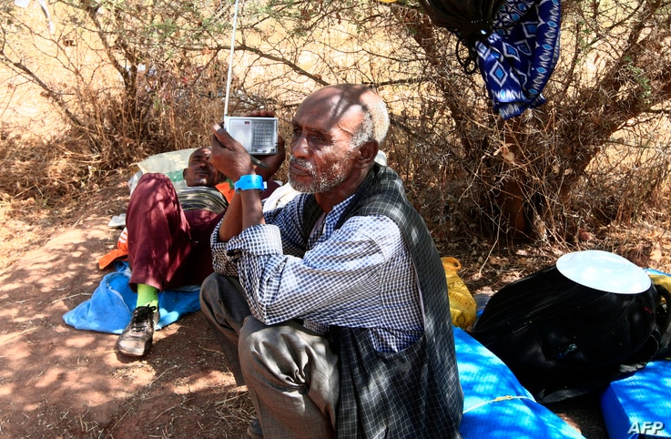 An Ethiopian refugee who fled fighting in Tigray province sits holding a radio in the shade of a straw shack at the Um Raquba…