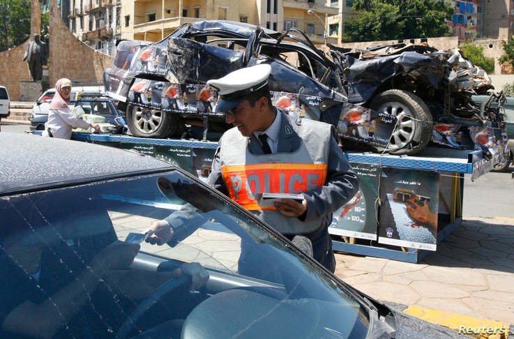 A policeman passes out leaflets with road accident statistics, in front of a destroyed car which is put on display, during a…